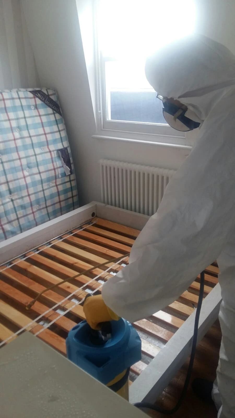 A BuzzKill technician treating a bed frame with residual insecticide. BuzzKill pest control technician in PPE applying insecticide to a bed frame during bed bug treatment